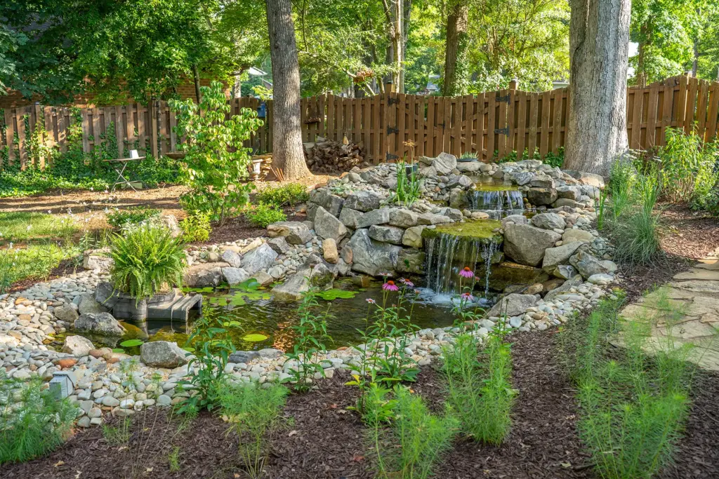 Landscape design Greenville SC: naturalistic backyard water feature with small pond, cascading waterfall over natural stone, water lilies, coneflowers, and surrounding native plantings